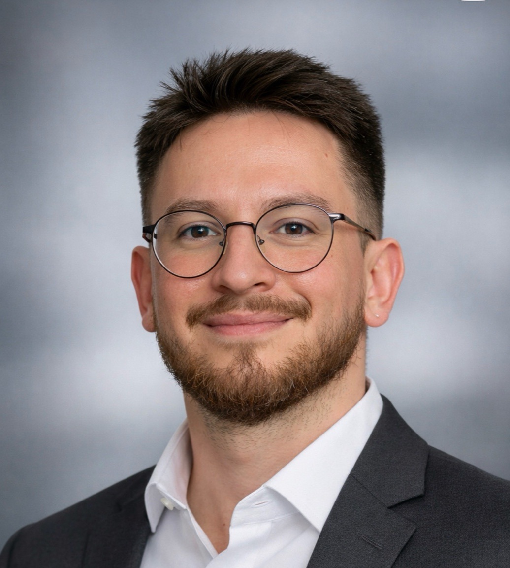 Head-and-shoulders portrait of a smiling man with short dark hair, round glasses, and a neatly trimmed beard, wearing a white shirt and dark blazer against a gray backdrop. | Akrual Accounting | Akrual Accounting